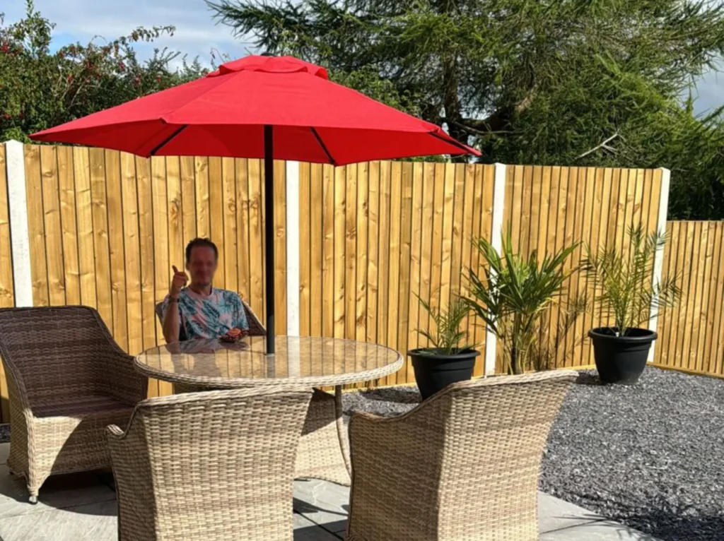 Person seated at a glass outdoor table with wicker chairs, holding a drink. A red umbrella provides shade. Wooden fence and plants in the background.